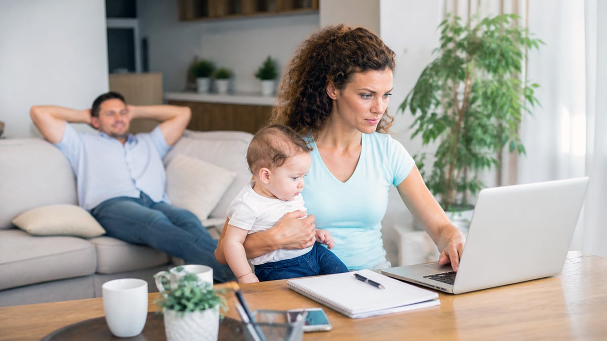 Mother working on a laptop while holding her baby in a bright living room as her partner relaxes nearby.